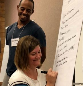 Woman smiling writing on whiteboard with a man smiling and standing next to her.