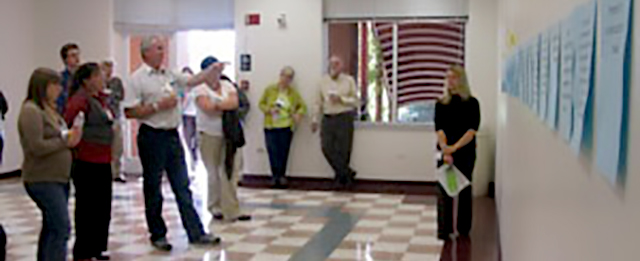 A group of people standing in a row, pointing at a wall with posters and talking.